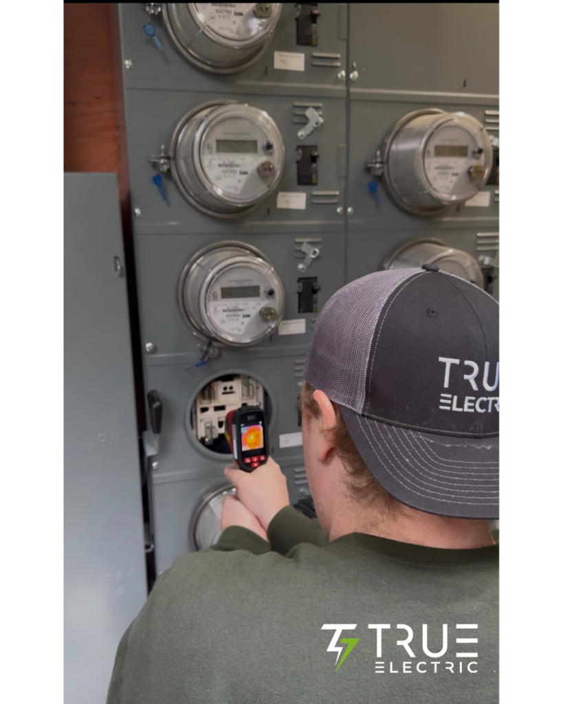 A licensed local electrician using a handheld infrared camera to perform an electrical thermal imaging scan on a building breaker panel.